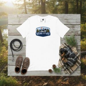 A white short-sleeve unisex t-shirt featuring the "Weenie in the Wild" graphic: a dachshund silhouette against a minimalist mountain range and retro sunset.