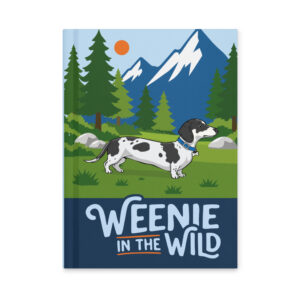 Front cover of the Weenie in the Wild matte hardcover journal featuring a black and white dapple dachshund in a mountain forest landscape.