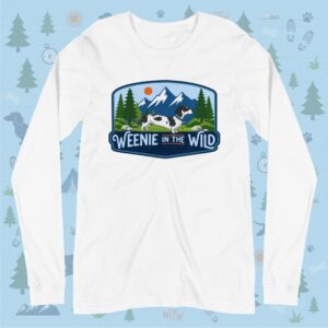 Front layout view of a solid white unisex long-sleeve crewneck tee shirt. The piece lies flat against a light blue outdoor decorative background, showcasing the vibrant "Weenie in the Wild" mountain and dapple dachshund logo centered on the chest.